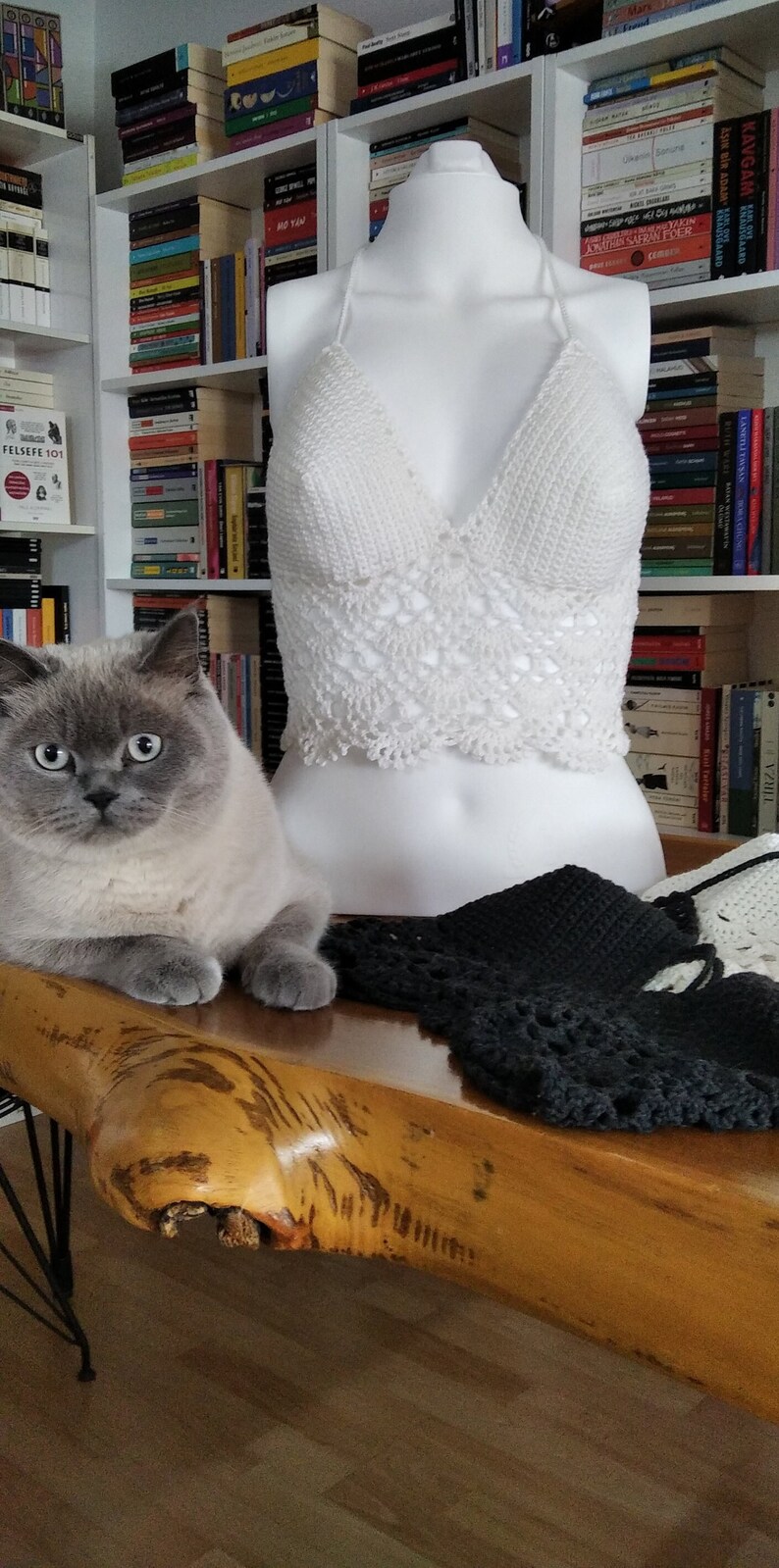 Pode incluir: Um top branco de croch&ecirc; com al&ccedil;as e um padr&atilde;o delicado, exibido em um manequim branco. O top est&aacute; sobre uma mesa de madeira com um gato cinza sentado na mesa.
