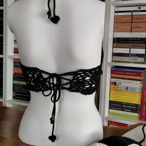 Pode incluir: Um top halter de croch&ecirc; preto com amarra&ccedil;&atilde;o nas costas e borlas pretas. O top &eacute; exibido em um manequim branco.