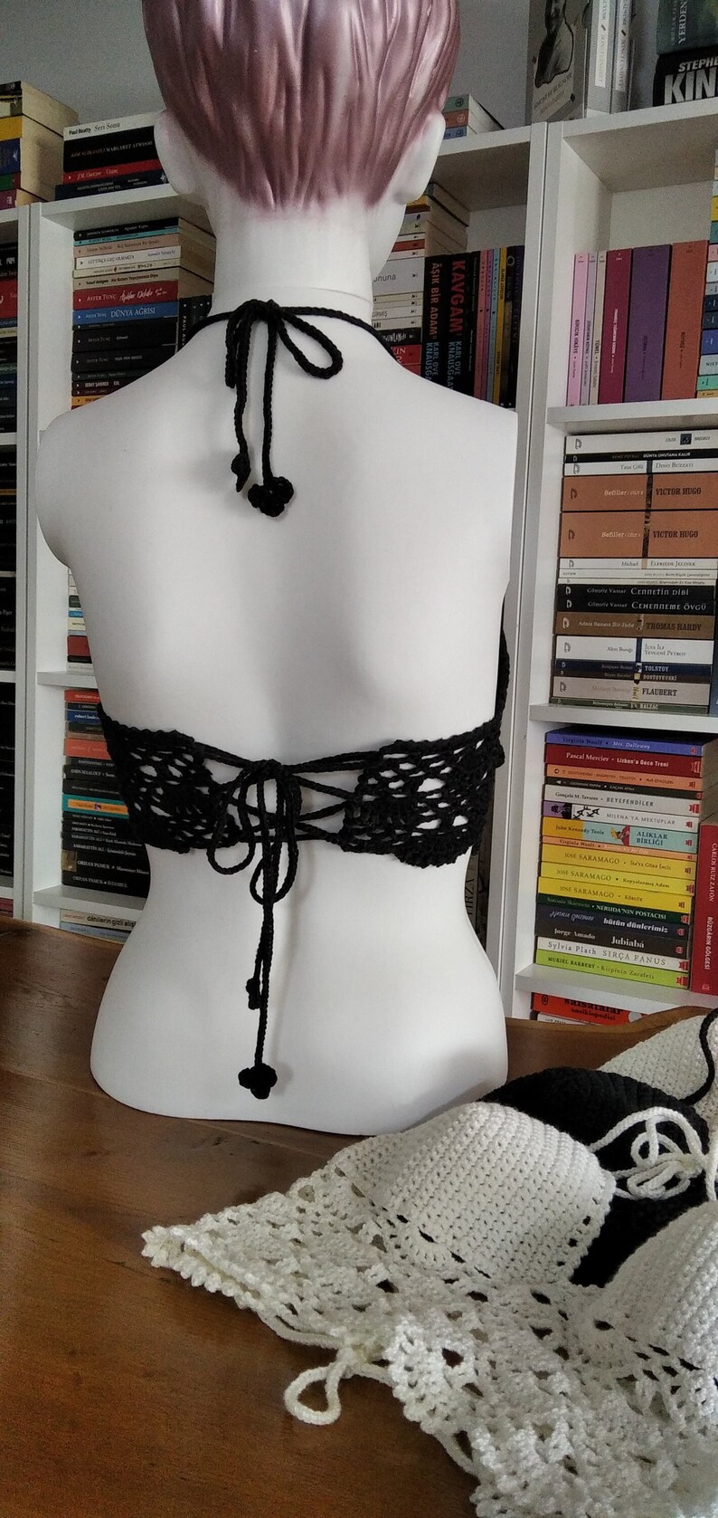 Pode incluir: Um top halter de croch&ecirc; preto com amarra&ccedil;&atilde;o nas costas e borlas pretas. O top &eacute; exibido em um manequim branco.
