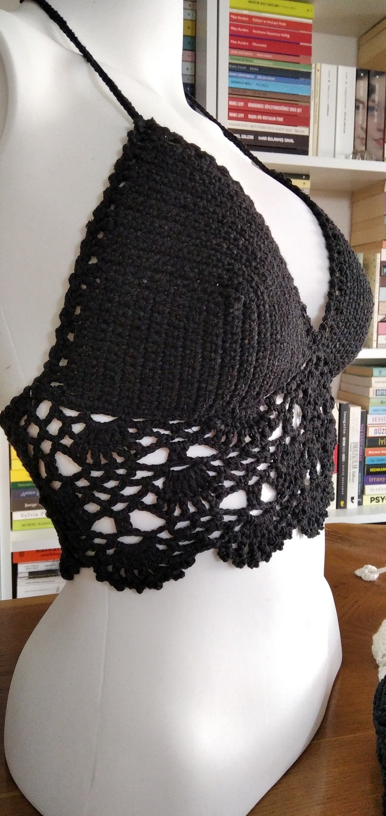 Pode incluir: Um top halter preto de croch&ecirc; com uma delicada borda de renda. O top &eacute; mostrado em um manequim branco.