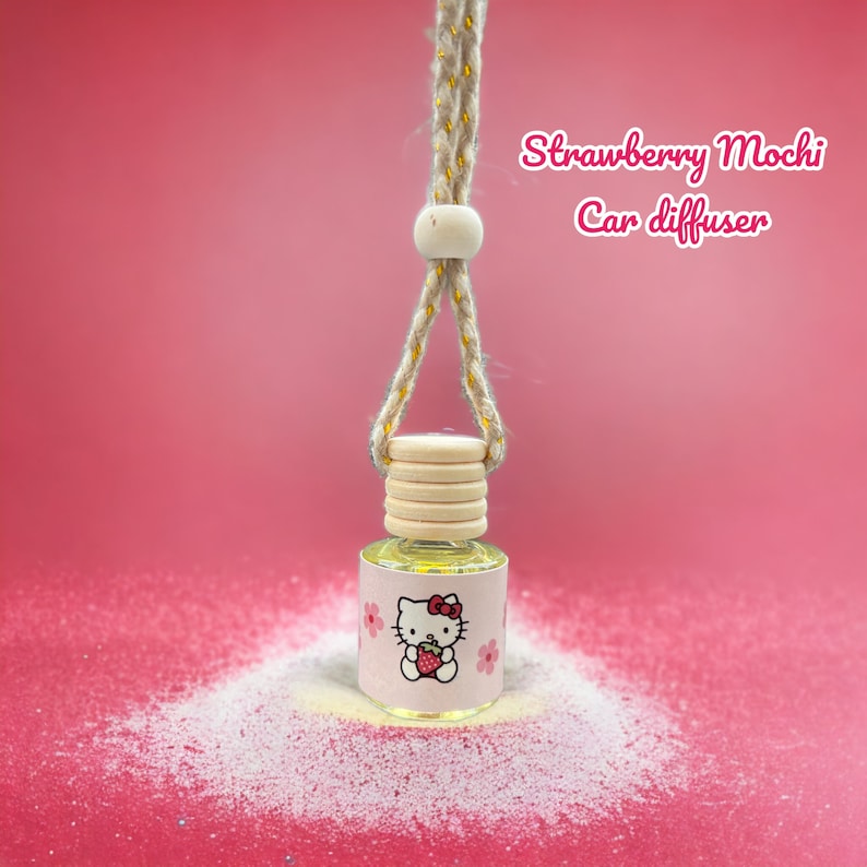 Pode incluir: Um ambientador para carro com tampa de madeira e cord&atilde;o para pendurar. O r&oacute;tulo apresenta um gato de desenho animado segurando um morango, com o texto "Strawberry Mochi Car diffuser" em rosa. O fundo &eacute; rosa.