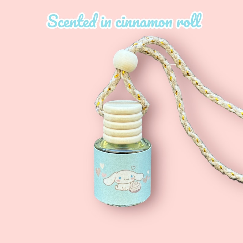 Pode incluir: Uma pequena garrafa de vidro com um r&oacute;tulo azul claro com um personagem de desenho animado e cora&ccedil;&otilde;es. A garrafa &eacute; coberta com uma tampa de madeira e uma corda bege. O texto na imagem diz "Scented in cinnamon roll".