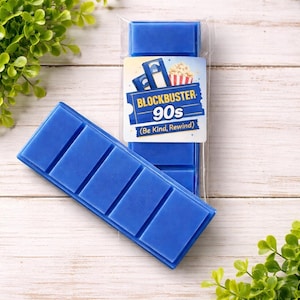 May include: Blue wax melt bars with a Blockbuster 90s label. The label features a VHS tape, popcorn, and the text "(Be Kind, Rewind)". The wax melts are arranged on a light wood surface with green foliage.