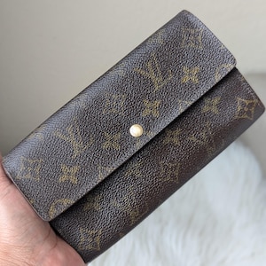 May include: Brown wallet with a flap closure and a gold-tone button. The wallet features a repeating pattern of interlocking logos. The leather wallet is held in a hand.