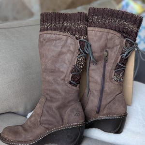 May include: A pair of brown UGG boots with a wedge heel. The boots have a knit cuff at the top and a zipper on the side. The boots have a leather upper and a rubber sole. The boots are approximately 12 inches tall.