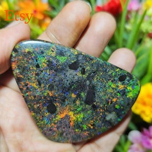 May include: A polished black opal gemstone with vibrant flashes of green, blue, yellow, and orange. The opal has a dark base colour with a speckled pattern and several dark spots. The word "Etsy" is visible in the upper left corner.