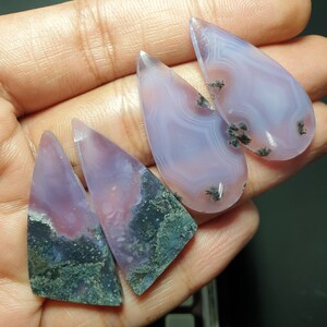 Puede incluir: Cuatro cabujones de ágata en tonos de morado, rosa y blanco con inclusiones negras y verdes. Los cabujones están cortados en forma de lágrima y triangular.