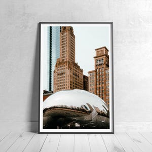 May include: Framed photograph featuring a polished, reflective, silver sculpture in the foreground, with several tall, brown brick buildings and a blue glass skyscraper in the background. The artwork is displayed on a white wooden floor.