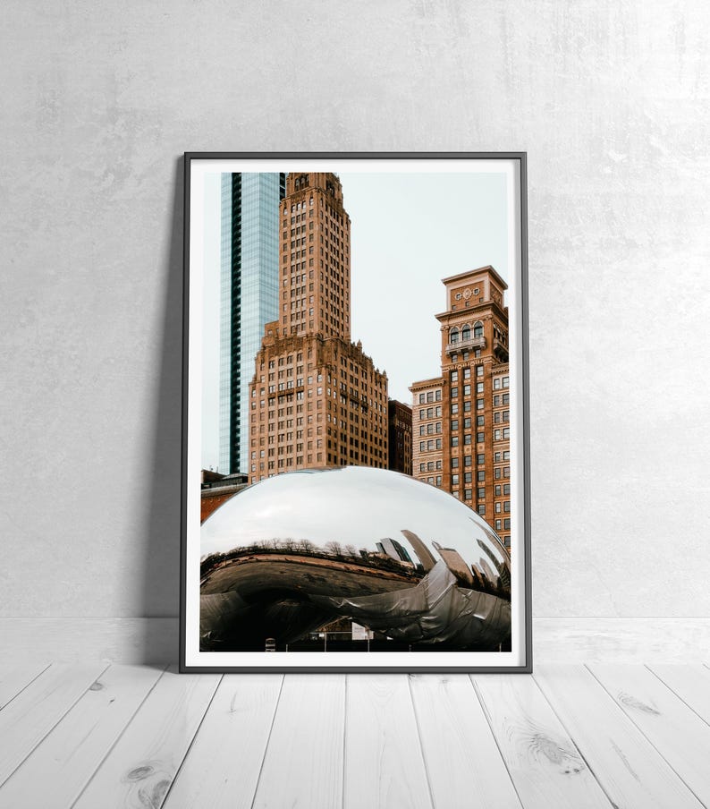 May include: Framed photograph featuring a polished, reflective, silver sculpture in the foreground, with several tall, brown brick buildings and a blue glass skyscraper in the background. The artwork is displayed on a white wooden floor.
