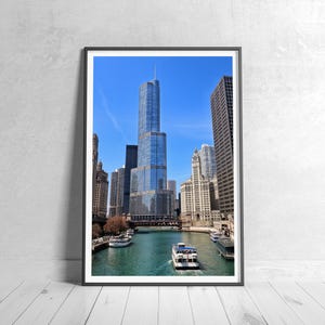 May include: Framed print of a cityscape featuring a tall, modern skyscraper with a glass facade, set against a clear blue sky. The image includes a river with boats, surrounded by other buildings. The artwork is displayed on a white wooden floor.
