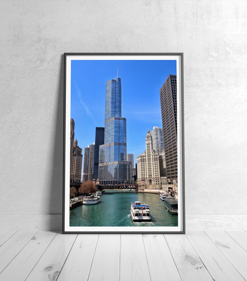 May include: Framed print of a cityscape featuring a tall, modern skyscraper with a glass facade, set against a clear blue sky. The image includes a river with boats, surrounded by other buildings. The artwork is displayed on a white wooden floor.