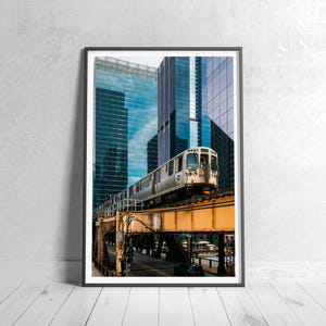May include: Framed photograph of a city train on an elevated track. The train is silver and white, with a weathered appearance. The track is supported by a yellow metal structure. Tall, modern buildings with reflective glass facades form the backdrop.