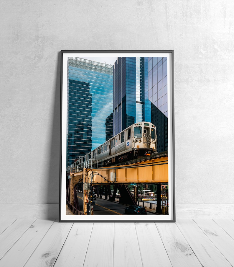 May include: Framed photograph of a city train on an elevated track. The train is silver and white, with a weathered appearance. The track is supported by a yellow metal structure. Tall, modern buildings with reflective glass facades form the backdrop.
