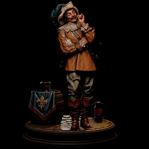 Puede incluir: Una figura detallada de un mosquetero con jubón color canela, sombrero de ala ancha con pluma azul y botas altas marrones. La figura se alza sobre una base de adoquines junto a un barril de madera cubierto con una tela azul con una flor de lis dorada.