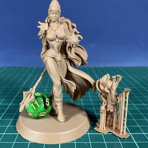 May include: A beige fantasy figurine of a woman in a flowing gown, crown, and cape, holding a staff and a small object. A green and white die is next to the figurine. A smaller, separate figurine is also present.