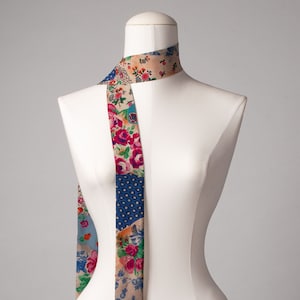 Könnte beinhalten: Ein langer, dünner Schal mit Patchwork-Muster, das florale Muster in Rosa-, Blau- und Grüntönen auf beigefarbenem Hintergrund zeigt.