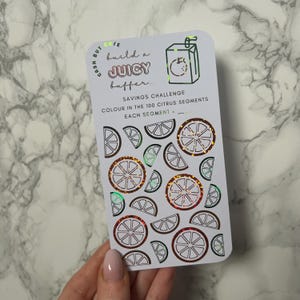 May include: A white savings challenge card with a citrus theme. The card features illustrations of orange slices and the text "build a JUICY buffer..." and "SAVINGS CHALLENGE COLOUR IN THE 100 CITRUS SEGMENTS". The card has a holographic foil effect.