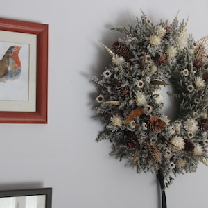 Op de afbeelding: Een witte en bruine gedroogde bloemenkrans met dennenappels en een roodborstje-illustratie in een houten lijst.