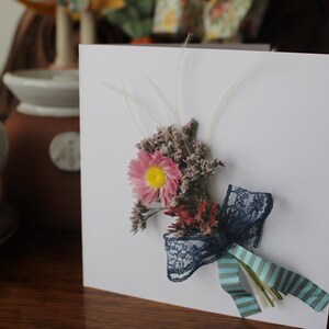 A mini bouquet of pinks, purples and feathery grasses with a blue lace and blue stripped ribbon on a gift card.