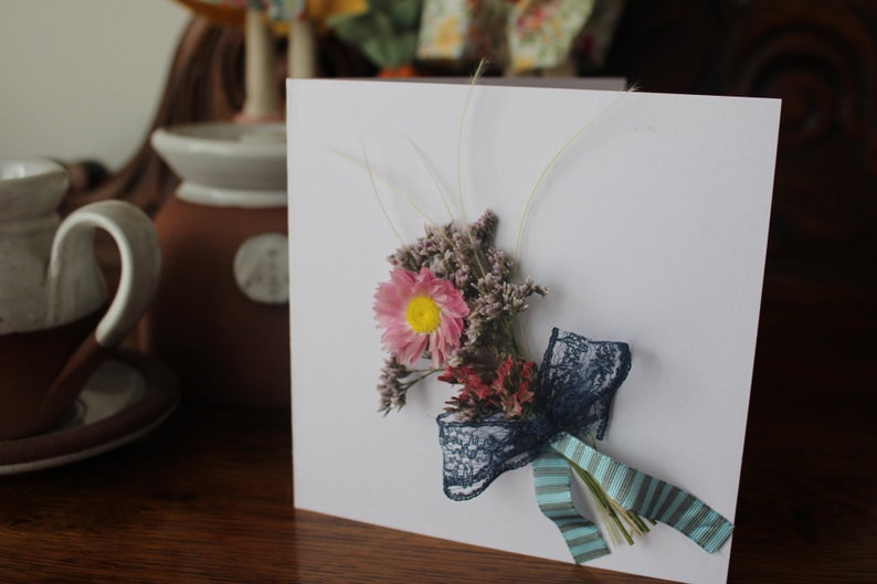 A mini bouquet of pinks, purples and feathery grasses with a blue lace and blue stripped ribbon on a gift card.