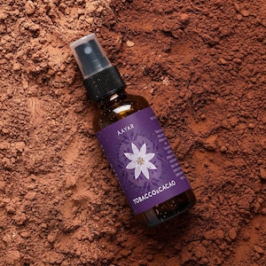 May include: A brown glass spray bottle with a black cap and a purple label. The label features a white floral design and the text "TOBACCO & CACAO". The bottle is set against a backdrop of brown powder.