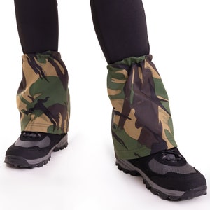 May include: A pair of black leggings with camouflage gaiters worn over the legs. The gaiters are made of a fabric with a green, brown, and black camouflage pattern. The person is wearing black hiking boots.