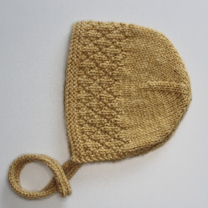 May include: A knitted baby bonnet in a light beige colour with a tied string. The bonnet has a diamond pattern.