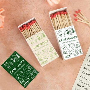 May include: Three matchboxes with red-tipped matches. The boxes are decorated with camping-themed illustrations and text. One box is green and says "Camp Lucas Bachelorette Weekend." Two boxes are off-white and say "Camp Riley" and "Camp Harper Bachelorette Weekend."
