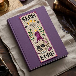 Cross stitch bookmark pattern on beige Aida with purple border: skull with a melting purple candle on top, wax drips, small flames, sprigs of flowers, stars, crescent moons, stitched heart, and text “SLOW BURN.” TourmalinePattern on Etsy.