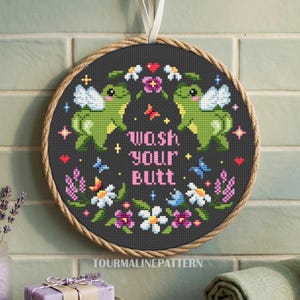 May include: A circular cross-stitch embroidery piece framed in a wooden hoop with a rope border. The dark gray fabric features two green frogs with white wings, colorful flowers, small butterflies, and the pink text "wash your butt" in the center.