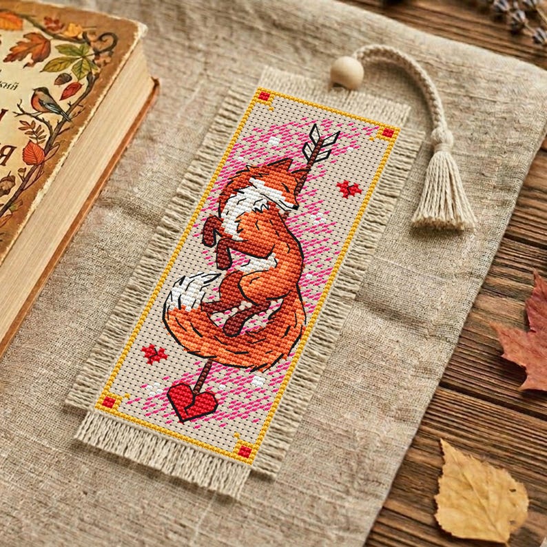Bookish cross stitch bookmark featuring a curled orange fox with arrow and red heart, displayed with an open book, TourmalinePattern on Etsy.