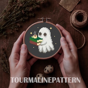 May include: A cross-stitch embroidery hoop featuring a white ghost holding a stack of books and a cup of coffee. The design is set against a dark gray background with yellow accents. The text "TOURMALINEPATTERN" is at the bottom.