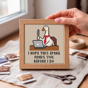 May include: A square cross-stitch embroidery in a light wood frame featuring a white goose wearing a red necktie, sitting at a brown desk with a grey laptop. The text below reads: I HOPE THIS EMAIL FINDS YOU BEFORE I DO. The design is on beige fabric.