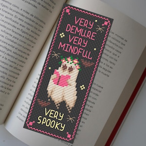 Op de afbeelding: Een zwart-witte bladwijzer met een roze geest die een boek leest. De geest draagt een roze bloemenkrans en de tekst "Very Demure Very Mindful Very Spooky" is in roze geschreven.
