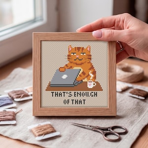 May include: A square wooden frame containing a cross-stitch embroidery of an orange tabby cat sitting at a desk with a laptop and a mug. The text below the cat reads, "THAT'S ENOUGH OF THAT." The embroidery is set against a light beige fabric background.