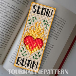 May include: Cross-stitch bookmark with the words "SLOW BURN" above and below a red heart engulfed in yellow flames. Green leaf accents are on either side of the flames. The bookmark is on a book.