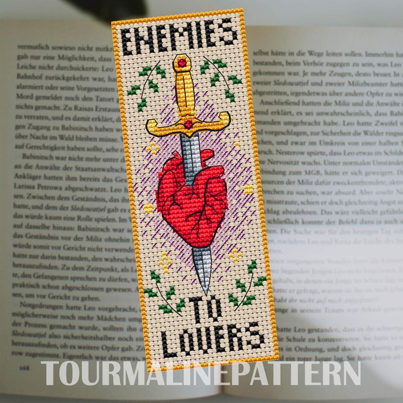Cross stitch bookmark featuring a red anatomical heart pierced by a golden sword, surrounded by decorative leaves and sparkles. The text 'Enemies to Lovers' is stitched in bold black lettering above and below the central motif.