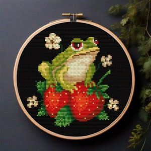 Puede incluir: Una rana verde con ojos rojos se sienta encima de dos fresas rojas con hojas verdes. La rana está rodeada de flores blancas. La imagen es un patrón de punto de cruz.