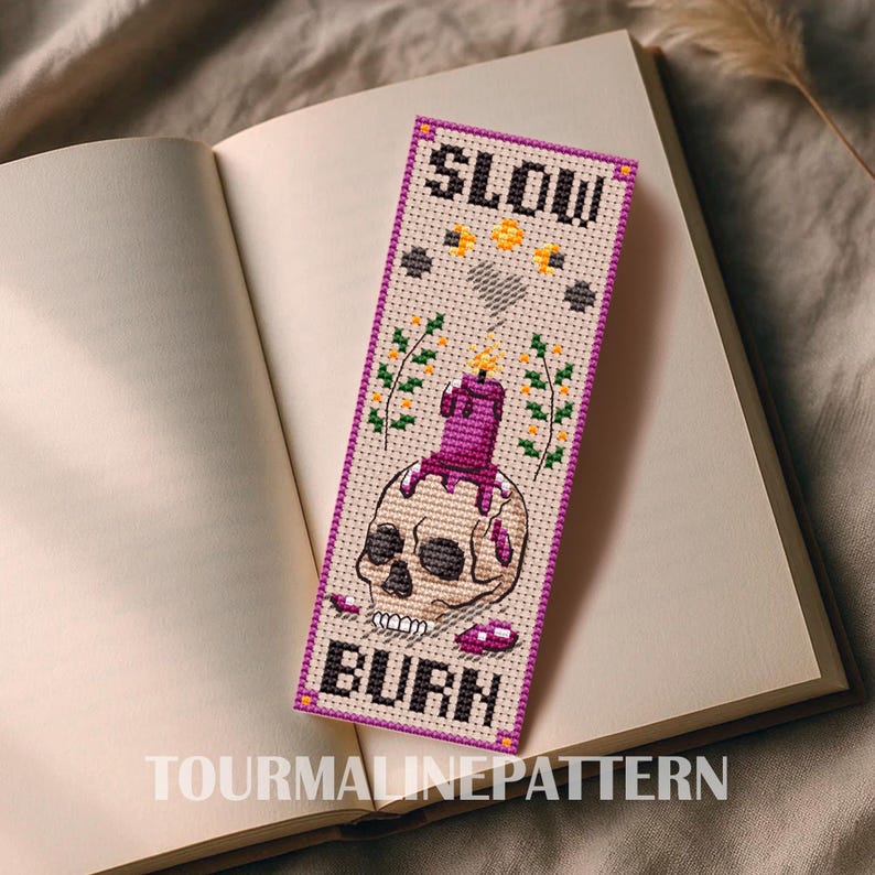Cross-stitch bookmark on beige Aida with a purple border: a skull topped with a melting purple candle, yellow sparks and flowers at the sides, tiny stars and motifs, and the text “SLOW BURN” at top and bottom. TourmalinePattern on Etsy