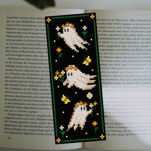 May include: A black bookmark with a green border features three white ghosts with orange flower crowns. The ghosts are holding flowers and are surrounded by yellow butterflies and white flowers.