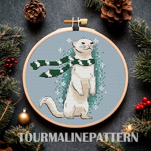May include: Cross stitch pattern of a white ferret wearing a green and white scarf on a blue background with white snowflakes.