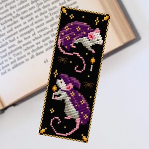 May include: A black bookmark with a gold border features two white mice wearing purple cloaks with gold stars. The mice are holding a candle and a star. The background is black with gold stars and spiders.
