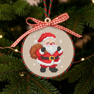 May include: A round Christmas ornament featuring a cross-stitch design of Santa Claus in his red suit, carrying a sack. The ornament has a red and green border, a red and white checkered ribbon, and a wooden bead for hanging. The ornament is hanging on a Christmas tree.