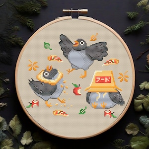 May include: Embroidered cross-stitch pattern featuring three cartoon birds wearing hats. The birds are surrounded by small details like leaves, apples, and pizza slices. The pattern is on a beige background and is framed in a wooden hoop.