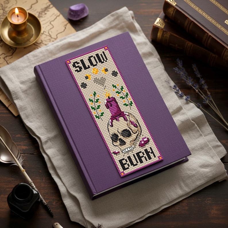 Cross stitch bookmark pattern on beige Aida with purple border: skull with a melting purple candle on top, wax drips, small flames, sprigs of flowers, stars, crescent moons, stitched heart, and text “SLOW BURN.” TourmalinePattern on Etsy.