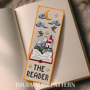 May include: Cross-stitched bookmark with a fantasy theme. The bookmark features a castle, mountains, dragons, clouds, and a crescent moon. The text "THE READER" is stitched at the bottom. The bookmark is placed on an open book.