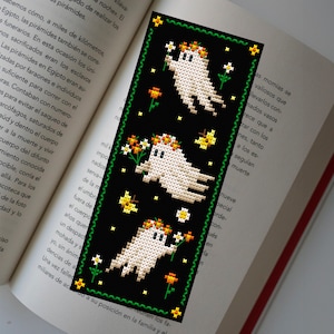 May include: A black bookmark with a green border features three white ghosts with orange and white flower crowns. The ghosts are holding white flowers and are surrounded by small white and orange flowers.