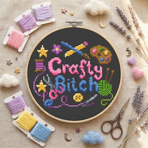 Peut inclure: Un tambour à broder circulaire en bois contenant un tissu noir avec un motif au point de croix. Le texte Crafty Bitch est brodé en lettres roses et bleues, entouré d'icônes de pinceau, palette, pelote, ciseaux et étoiles.