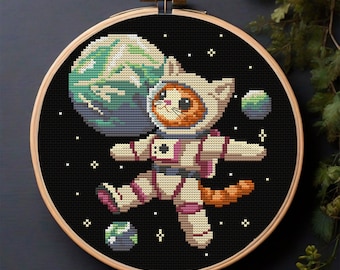 Puede incluir: Un aro de bordado de punto de cruz con fondo negro que presenta un gato de dibujos animados con un traje de astronauta blanco flotando en el espacio. El gato está rodeado de estrellas y planetas.