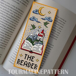 Fantasy cross-stitch bookmark: open book grows into mountains and a red-roof castle under a crescent moon, clouds, tiny stars, and two small dragons; “THE READER” text at the bottom, golden border. TourmalinePattern on Etsy